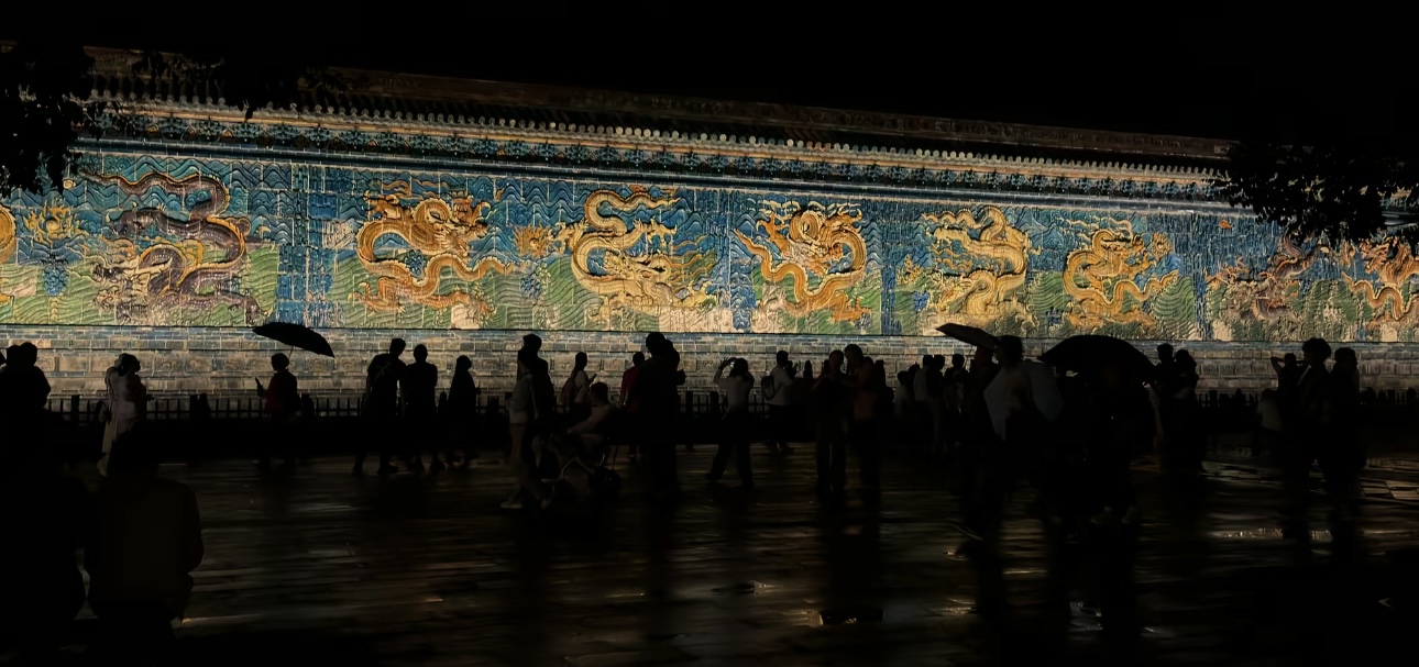 Visitors silhouetted at night in front of an illuminated glazed dragon screen